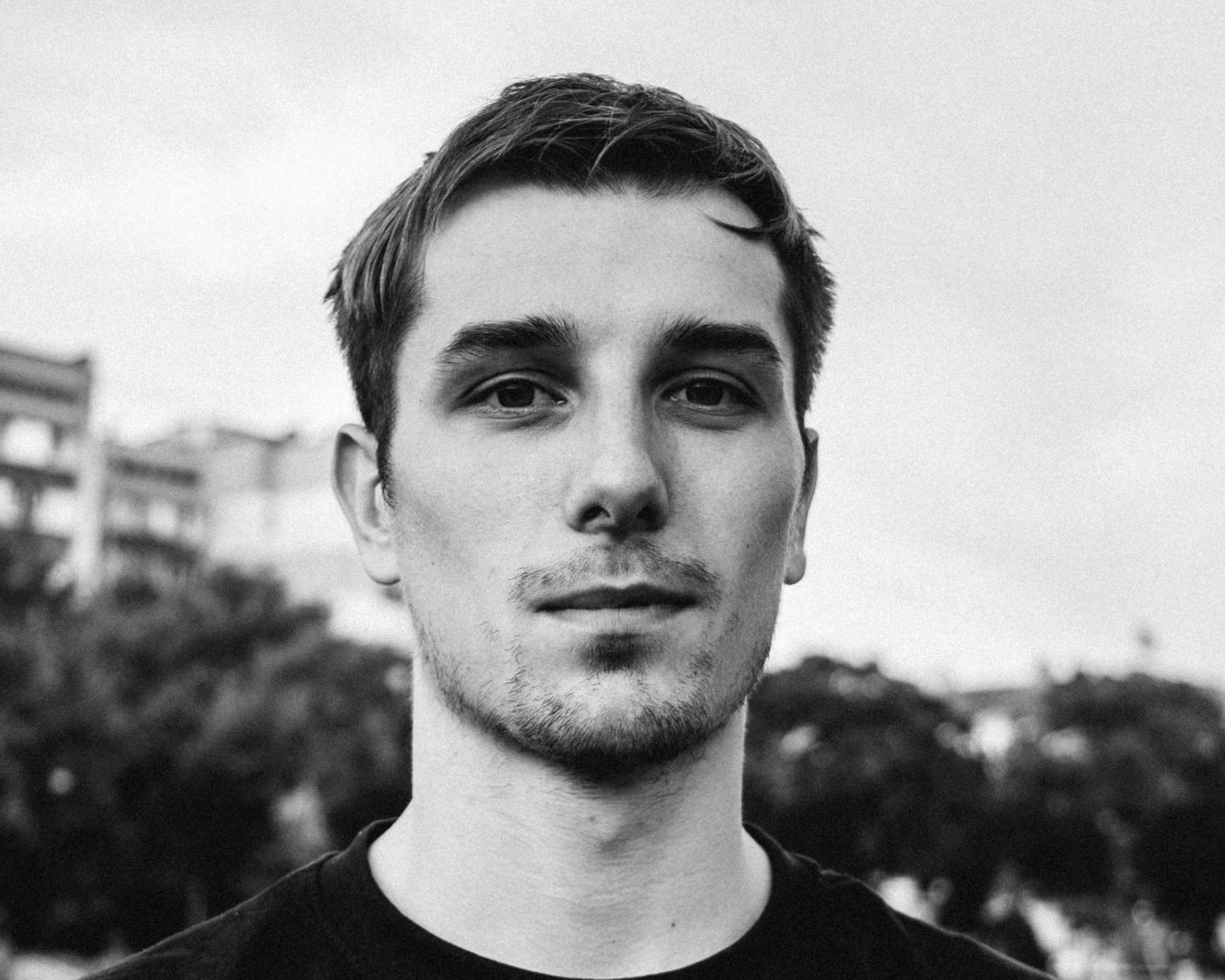 Black-and-white portrait of a young man with short hair and light facial stubble, looking directly at the camera with a neutral expression, outdoors with blurred buildings and trees in the background.